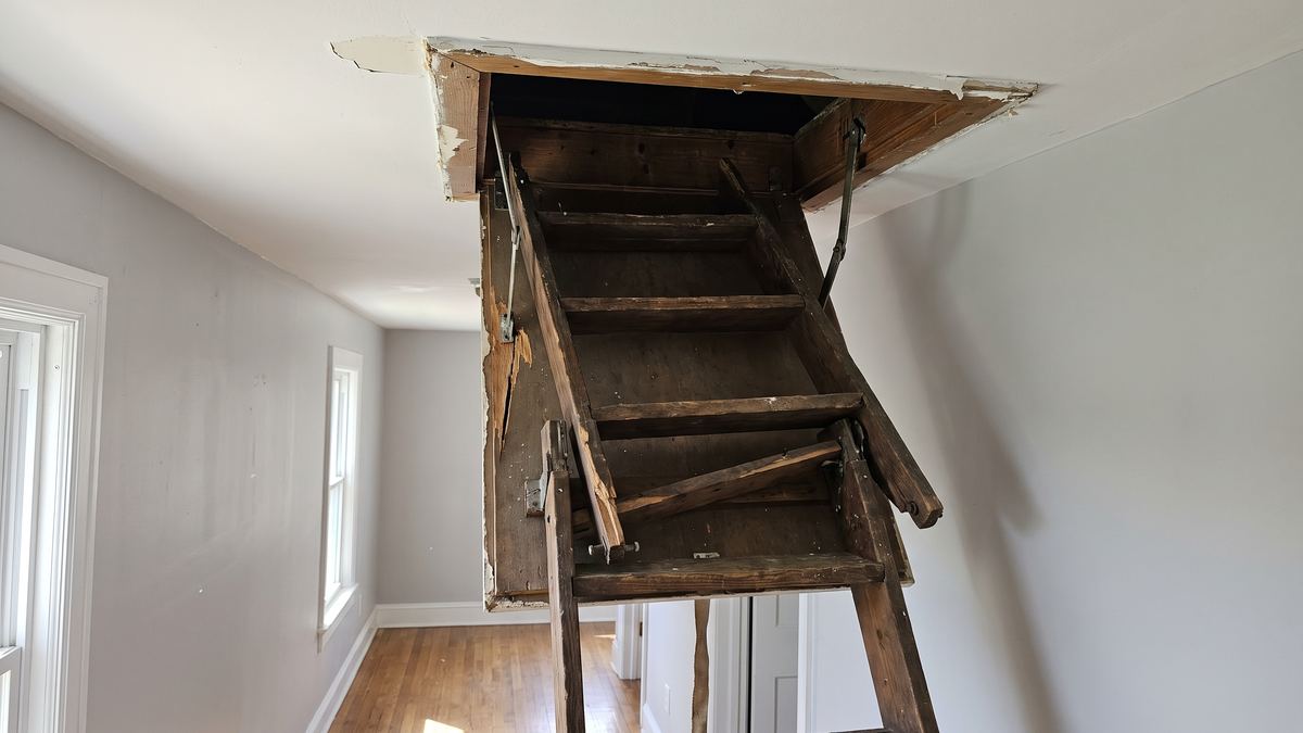 Hallway ceiling with a worn wooden attic ladder hanging crooked from its opening